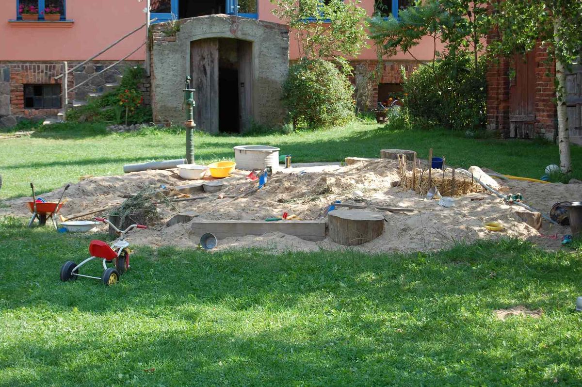 Sandfl&auml;che mit Spielzeug, Holzst&auml;mmen und einer gr&uuml;nen Handpumpe auf der Wiese