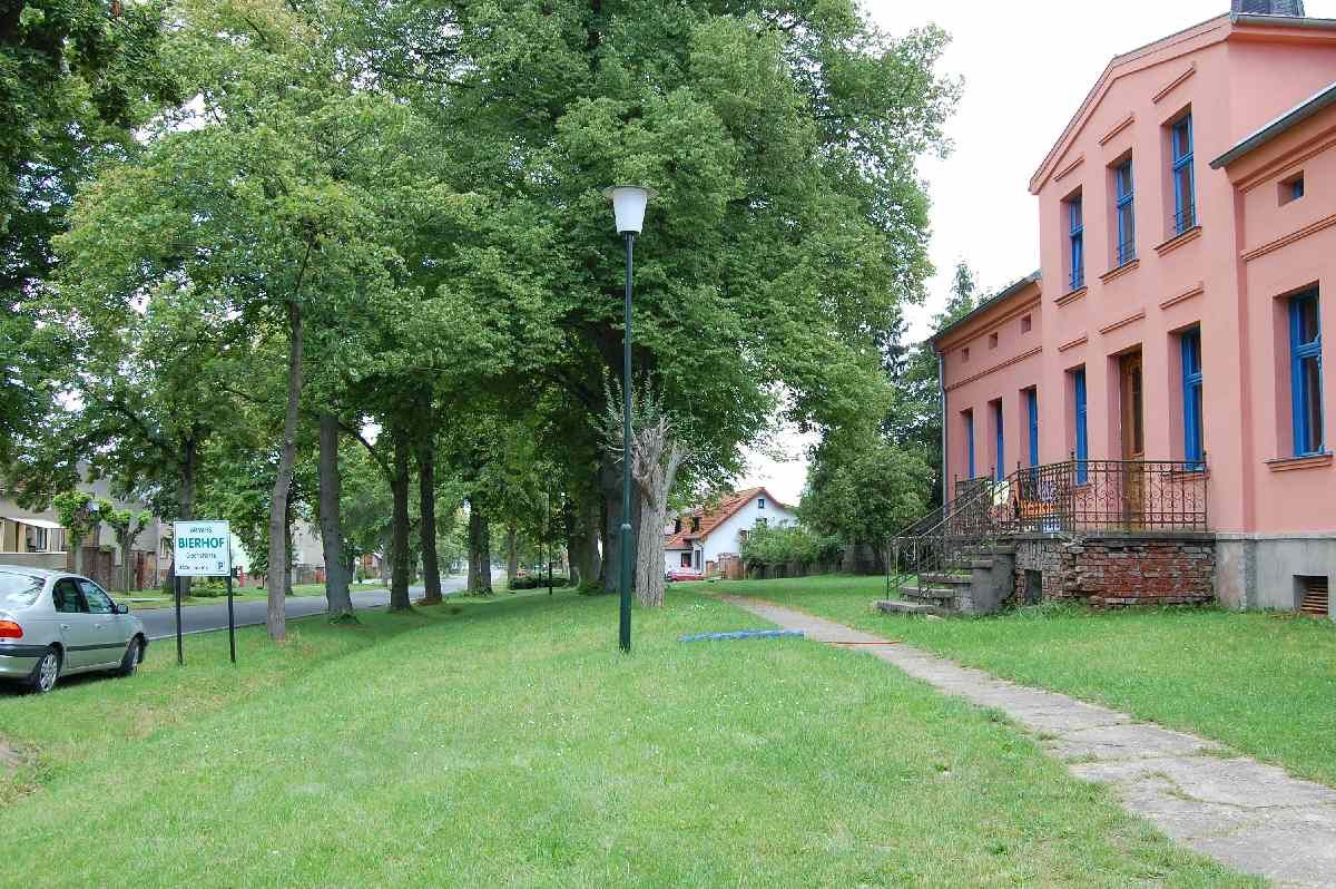 Dorfstra&szlig;e mit alten B&auml;umen, links ein Ortsschild, rechts ein rosa Haus mit blauen Fenstern